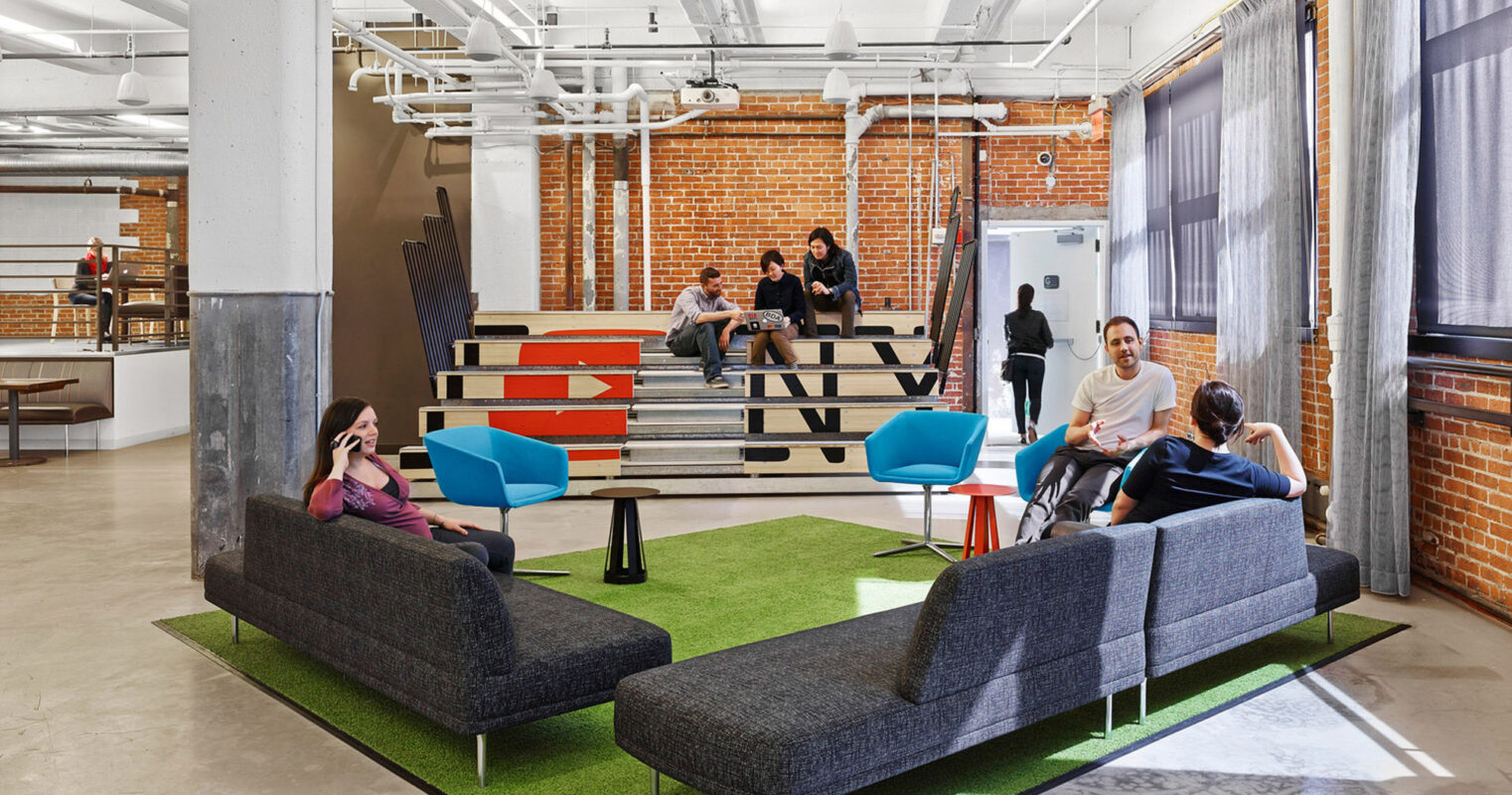 Modern loft-style office space with exposed brick walls and white ductwork against a high ceiling. Employees engage casually on colorful modular furniture atop a vibrant green area rug, reflecting an innovative and collaborative work environment.