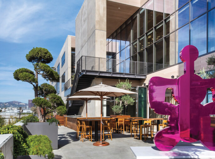 Rooftop terrace with modern outdoor furniture set against a backdrop of architectural glass and clean-lined structures. Purple abstract sculpture adds a pop of color, with greenery accentuating the urban landscape view.