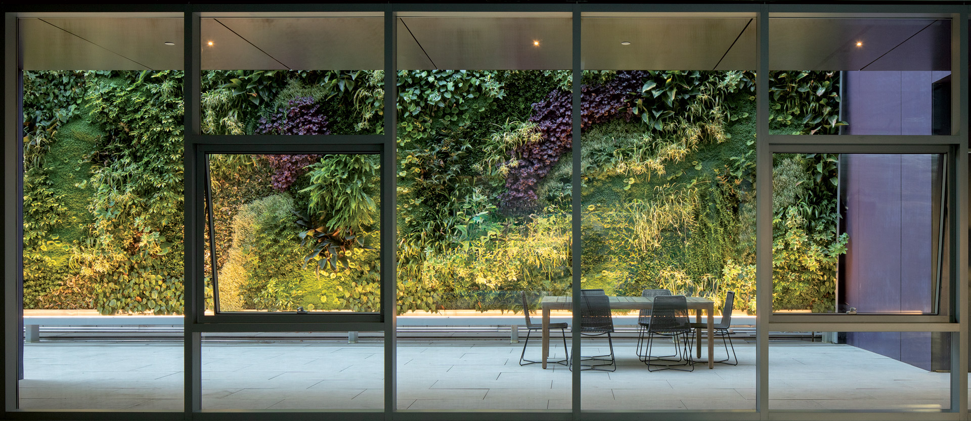 Spacious dining area with floor-to-ceiling glass walls, overlooking a vibrant vertical garden. Sleek furniture contrasts with lush greenery, under soft, ambient lighting for a natural, contemporary atmosphere.