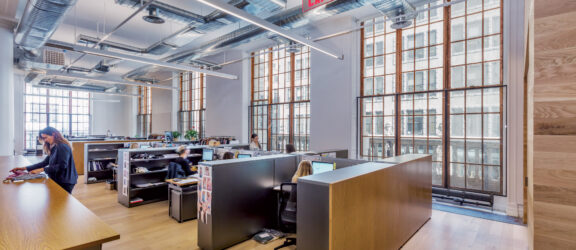 Open-plan office space with exposed ceiling infrastructure, large windows bathing the room in natural light, sleek black desks, and ergonomic chairs neatly organized, promoting a collaborative work environment.