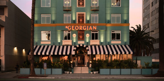Twilight bathes the façade of the Georgian Hotel, a symmetrical three-story building with a flat roof and a teal stucco exterior. It features contrasting white stone trim, decorative ironwork, and neon signage. The entrance is marked by a black and white striped awning, matching balcony canopies, and flanked by lush greenery.