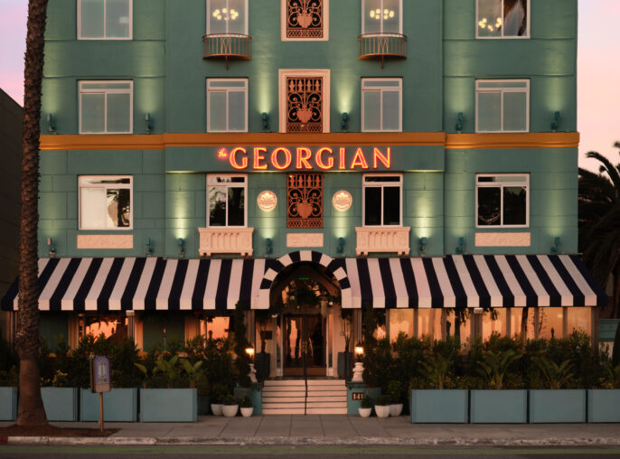 Twilight bathes the façade of the Georgian Hotel, a symmetrical three-story building with a flat roof and a teal stucco exterior. It features contrasting white stone trim, decorative ironwork, and neon signage. The entrance is marked by a black and white striped awning, matching balcony canopies, and flanked by lush greenery.