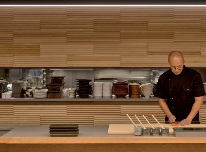 Chef meticulously prepares food at a sleek restaurant kitchen counter, framed by warm wooden slat wall panels that create a rhythmical texture, complemented by minimalist plateware and ambient lighting that casts a cozy glow over the culinary scene.