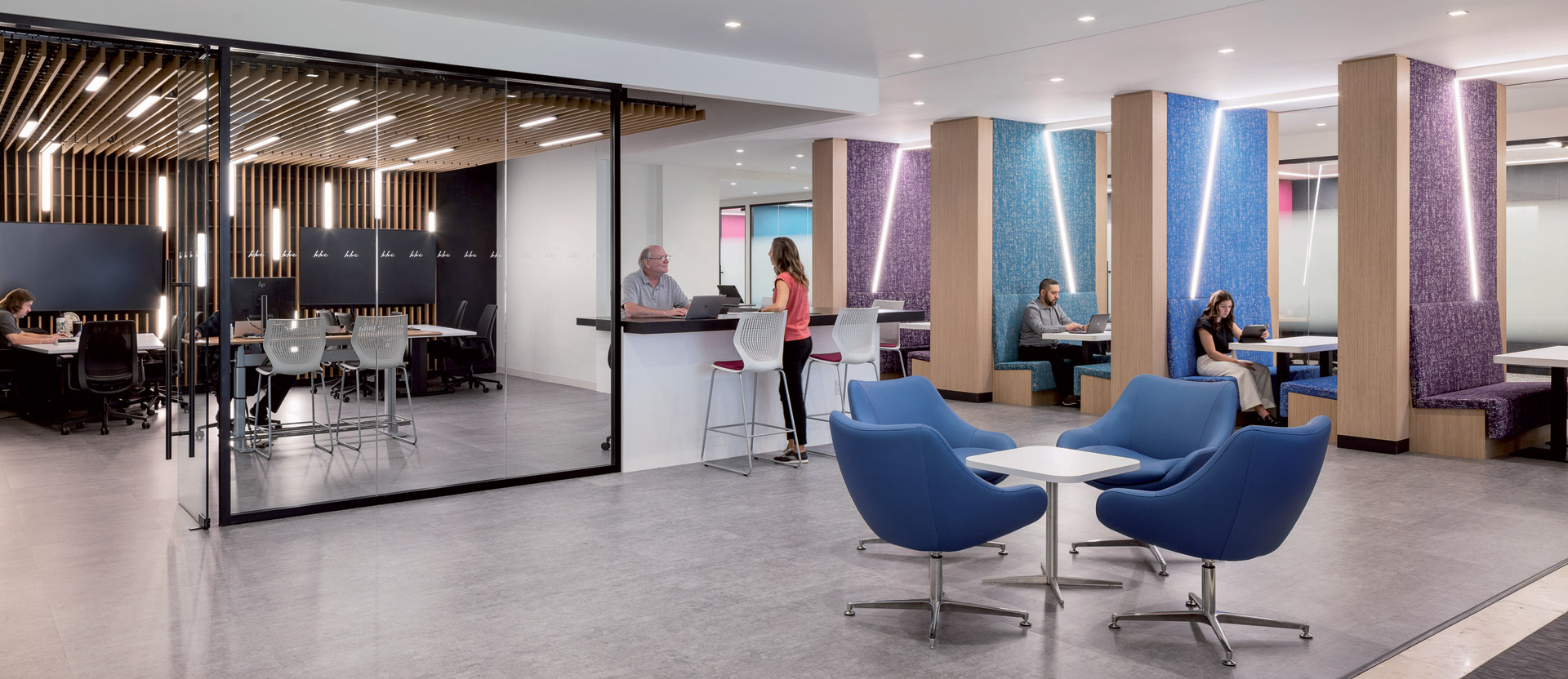 Contemporary office break room with vibrant blue egg chairs, sleek white high-top tables, and people in casual conversation. Textured columns with purple accents complement the neutral-toned flooring and wooden slatted ceiling design.