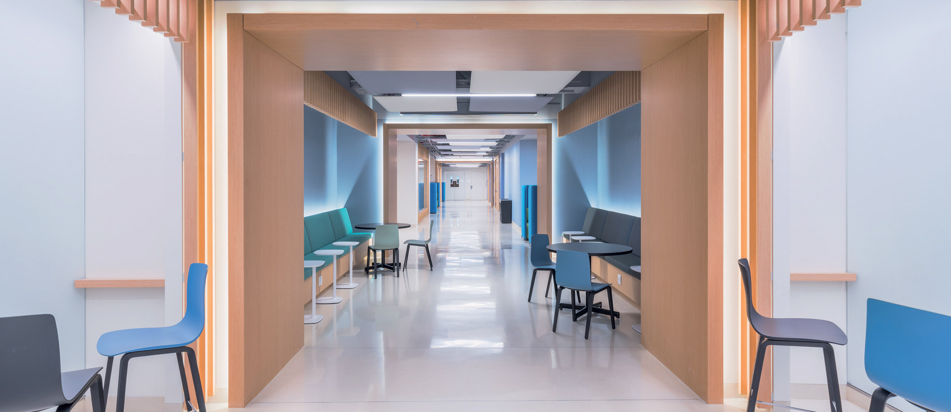 Modern corridor with clean lines, featuring wood-accented alcoves, teal and blue furnishing, and efficient LED lighting guiding the way. The design merges functionality with comforting aesthetics to ease navigation and enhance patient comfort.