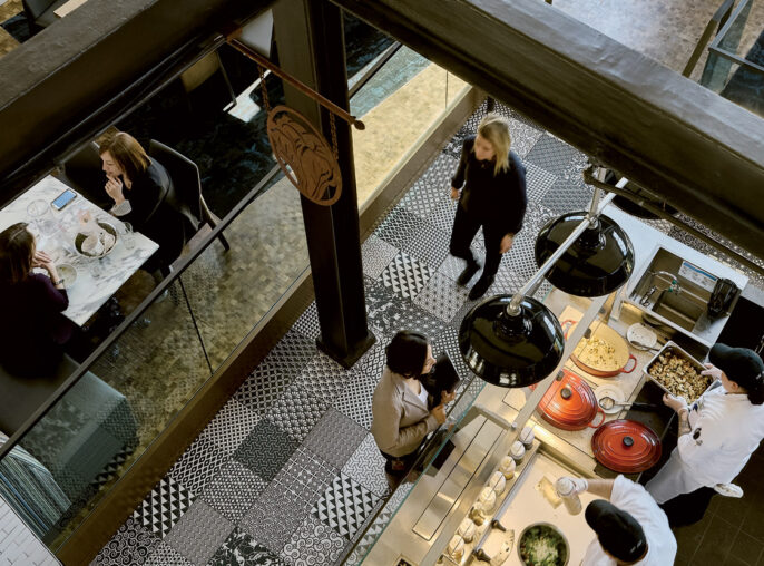 Overhead view of a bustling restaurant interior showing a contrast of textures with exposed brick walls, sleek wooden beams, geometric tiled flooring, and an eclectic mix of modern and rustic furniture; diners enjoy meals under warm, ambient lighting.