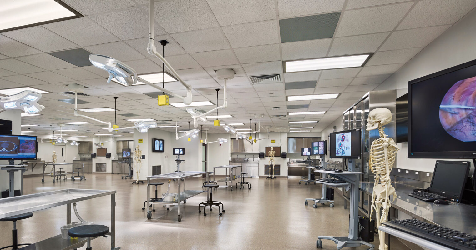 Spacious, modern medical laboratory interior with stainless steel autopsy tables, overhead surgical lights, and multiple computer stations. A human skeleton model stands on the right, with a large monitor displaying a cellular image in the background, reflecting a practical educational environment.