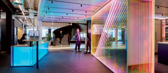 Modern office lobby with vibrant, multicolored LED lighting integrated into a slatted wooden feature wall. A sleek reception desk with ambient lighting complements the dynamic space, while contemporary furniture and ceiling details add to the sophisticated corporate aesthetic.