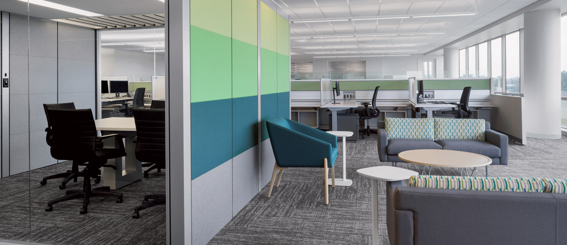 Open-plan office space featuring modular workstations with ergonomic chairs, individual privacy panels, and ambient natural lighting from floor-to-ceiling windows. A casual seating area introduces vibrant upholstery, complementing the space's contemporary, collaborative atmosphere.