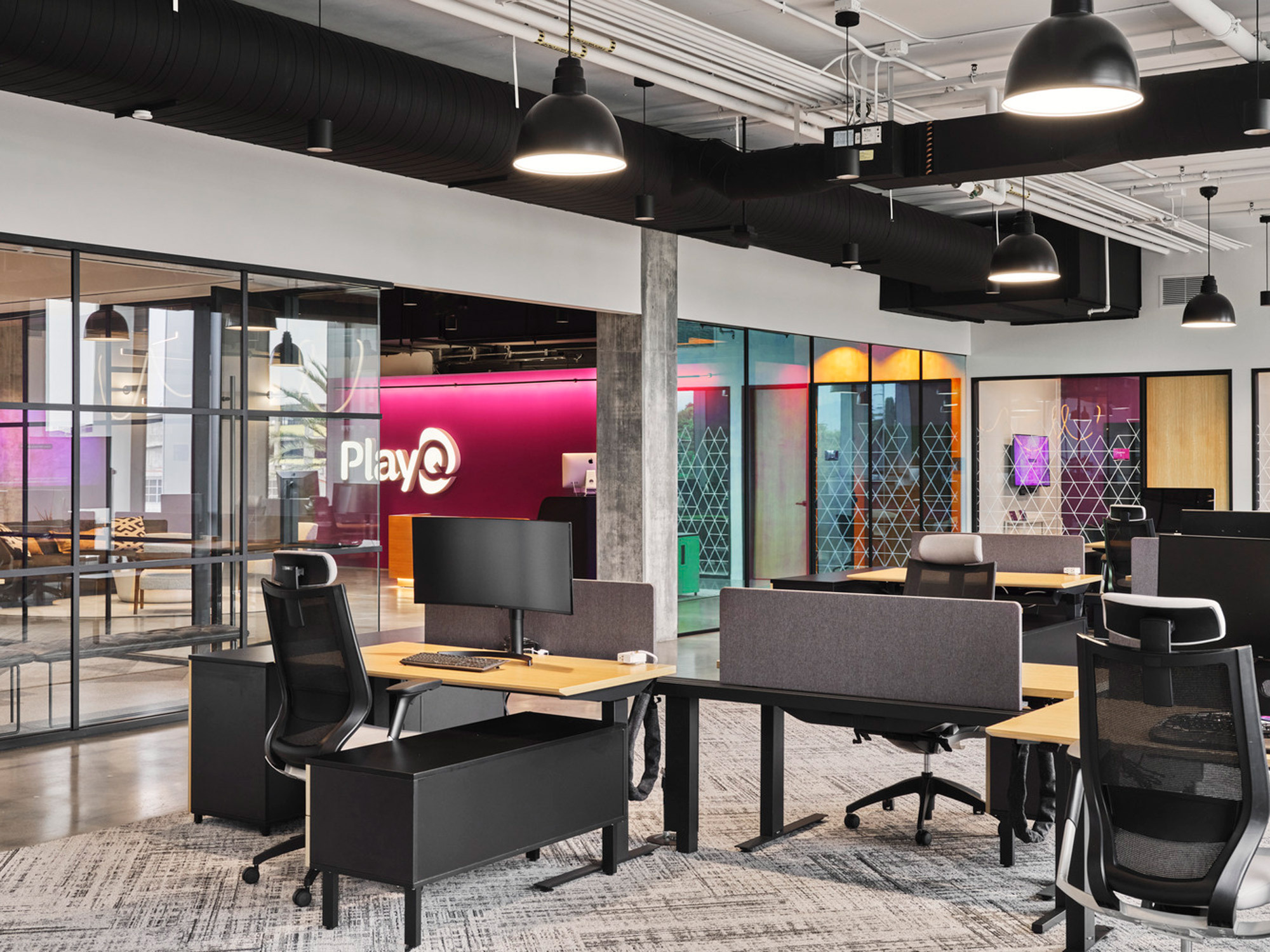 Modern office space featuring ergonomic chairs, sleek black desks, vibrant magenta branding wall, and glass partitions with playful designs contrasting with industrial exposed ceilings and ductwork.