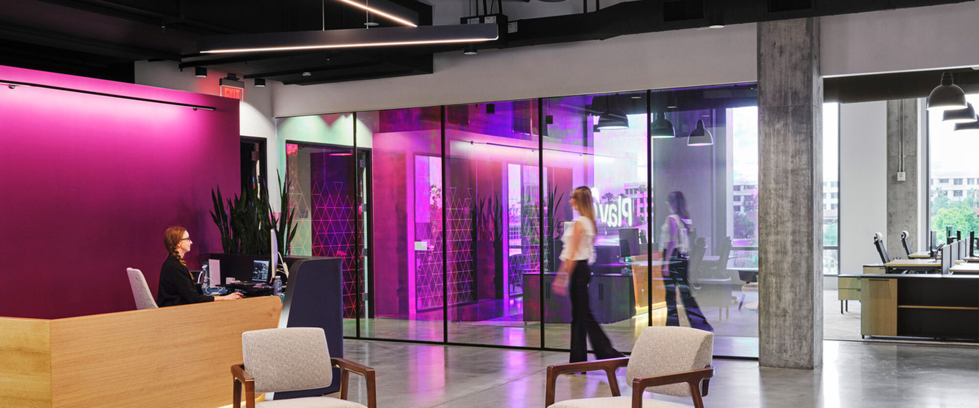 Modern office lobby with a minimalist reception desk, flanked by two white chairs. Vibrant magenta glass panels create a dynamic backdrop, while sleek, geometric light fixtures add a contemporary touch overhead. The design fosters an airy and welcoming atmosphere.