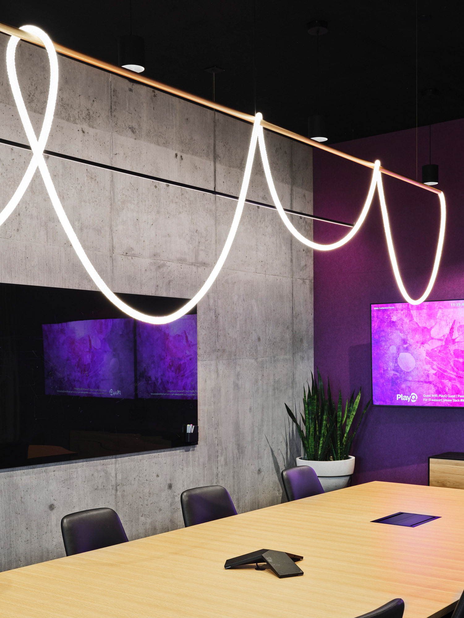 Modern conference room featuring a bold, organic LED lighting fixture above a wooden table, with concrete walls, integrated technology, and vibrant purple accent lighting adding a dynamic atmosphere.