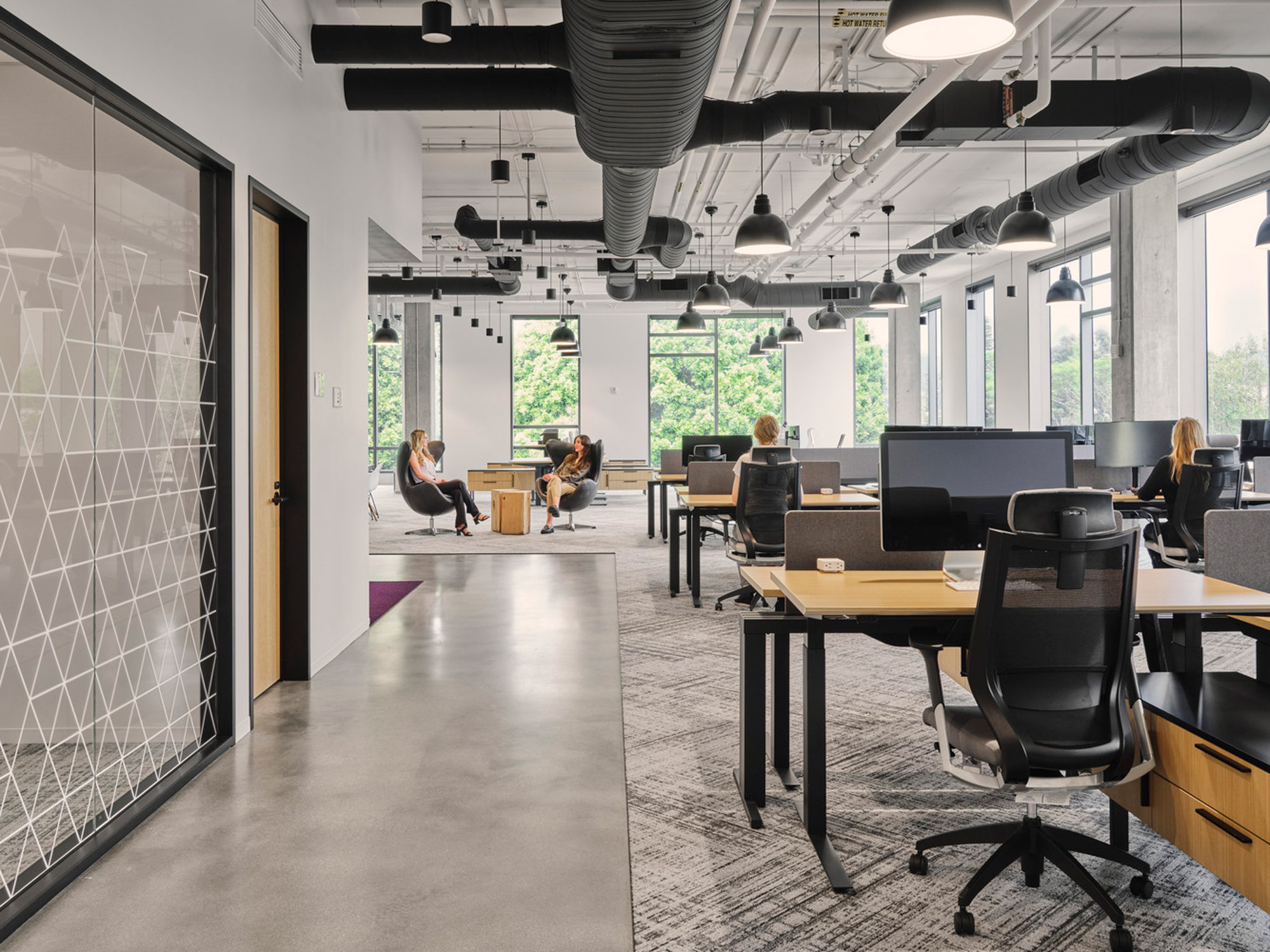 Modern office space with an open floor plan featuring exposed ductwork, sleek furnishings, and geometric glass partitions. Natural light permeates, illuminating the gray-patterned carpet and collaborative workstations.