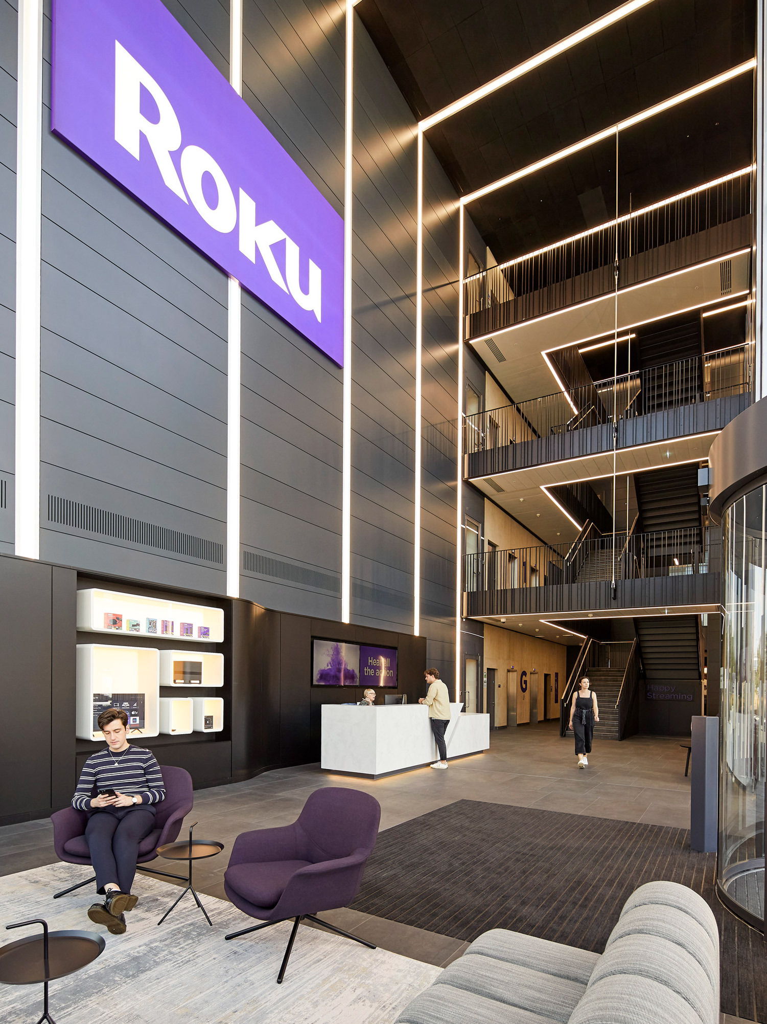 A modern corporate lobby featuring a tall ceiling with geometric black lines and recessed lighting. The Roku branding dominates the upper wall. Below, visitors navigate the open staircase while others sit on stylish purple chairs, surrounded by the warm textures of wood and gray carpeting.