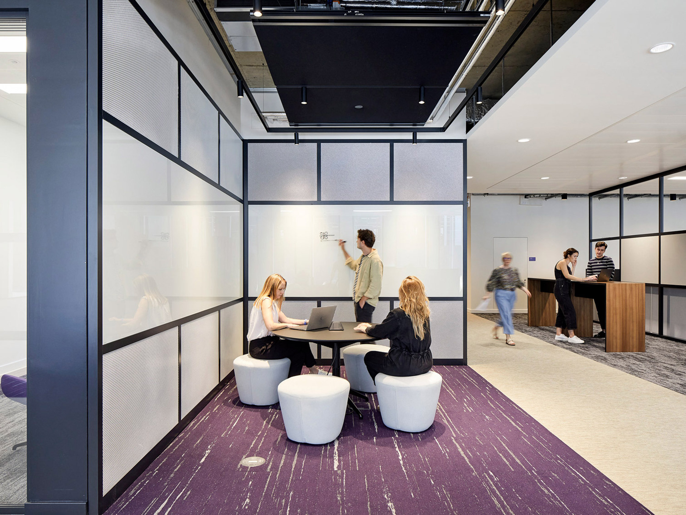 Modern office space featuring glass partitions with black frames, enhancing the open-concept layout. Whiteboard session in progress amidst plush white seating over a vibrant purple carpet, with ambient lighting and a wooden reception desk anchoring the background.