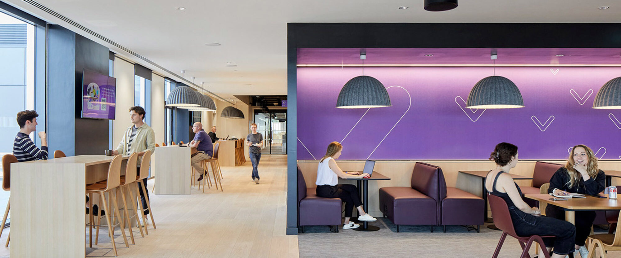 Modern office cafeteria with vibrant purple accent wall featuring playful line art, contrasted by natural wood tones and sleek furniture. Casual seating areas promote social interaction, complemented by ambient pendant lighting amidst an open, industrial ceiling design.
