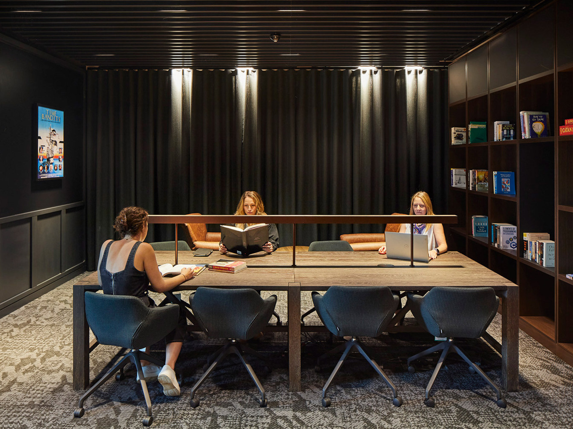 Modern corporate library space with individuals at a large shared wooden table, surrounded by dark shelving filled with books and accent lighting, creating an inviting atmosphere for collaboration or quiet study.