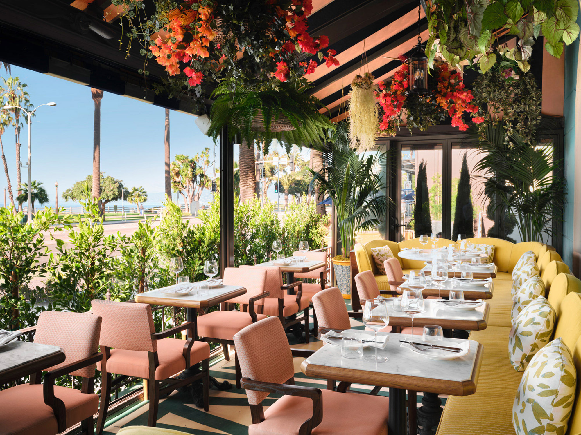 Open-air restaurant with a vibrant floral canopy, integrating nature with dining. Warm wood tones and pink chairs contrast against yellow banquette seating, capturing a breezy, coastal elegance. Ocean views complement the botanical ambiance, celebrating a seamless indoor-outdoor experience.