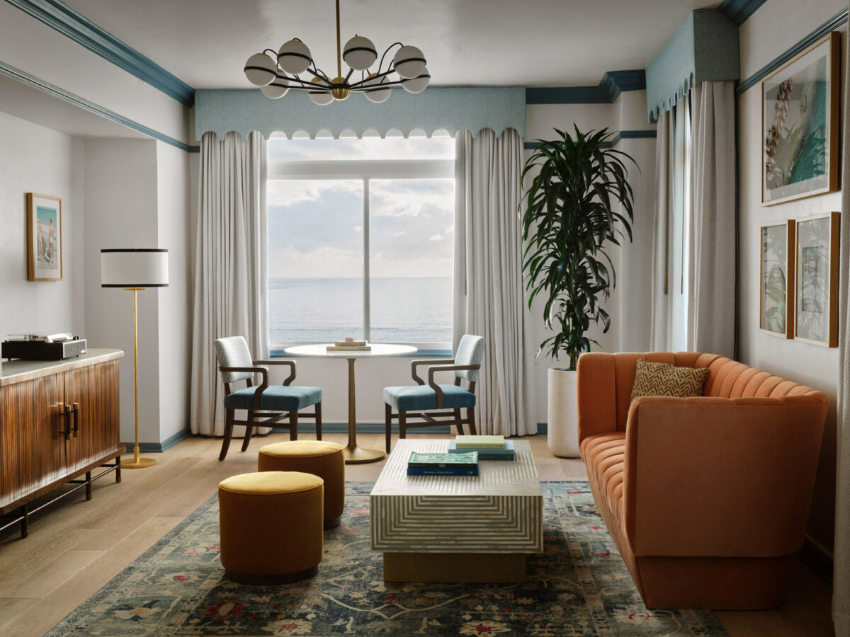 Coastal-inspired living space with expansive ocean view framed by floor-to-ceiling windows. Mid-century wooden sideboard, plush tufted sofas, and patterned area rug harmonize with muted color palette. Prominent indoor plant and retro chandelier accent the calm, sophisticated ambiance.