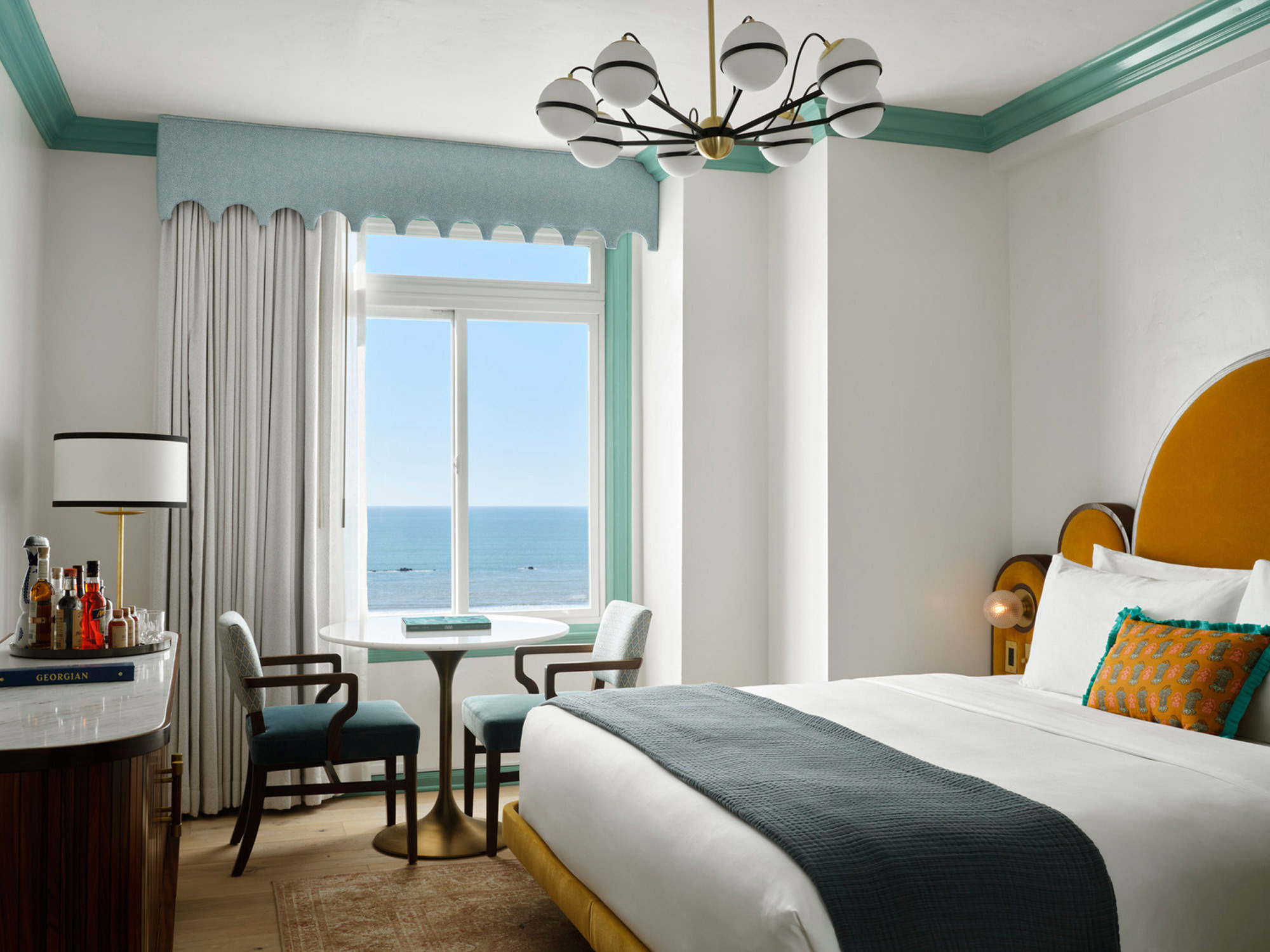 Oceanfront bedroom featuring a minimalist design with crisp white walls and a king-sized bed. Teal accents line the ceiling and window, complementing the marine view. A sculptural chandelier adds a modern touch above, while a round mirror and bespoke cabinetry offer functional elegance.