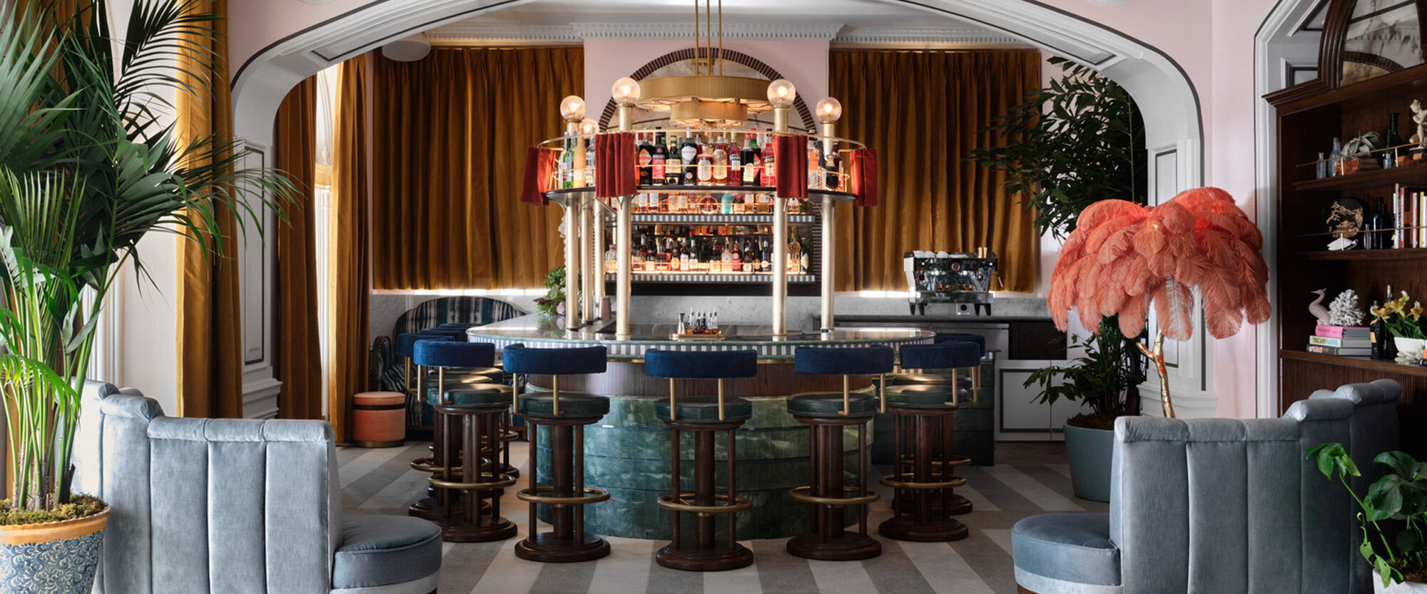 Elegant bar lounge featuring Art Deco style with a central circle bar, plush blue and grey seating, radiant floor patterns and an ornate gold chandelier providing a warm ambiance.