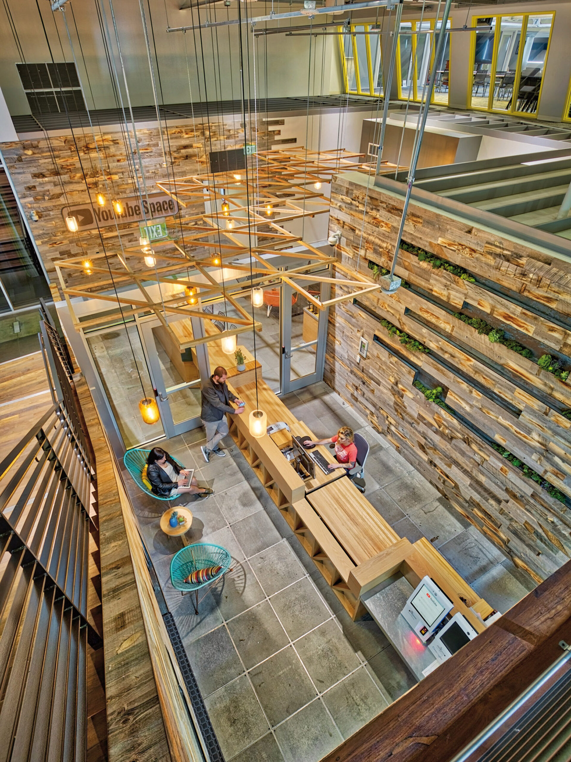 This modern atrium features an eclectic mix of industrial and natural design elements. Exposed steel beams support a network of suspended light fixtures, contributing to the loftiness of the space. Wood cladding, interspersed with greenery, creates a textured backdrop along one wall, while a terraced section offers informal seating. Polished concrete floors anchor the airy scheme, and furniture pieces with clean lines provide