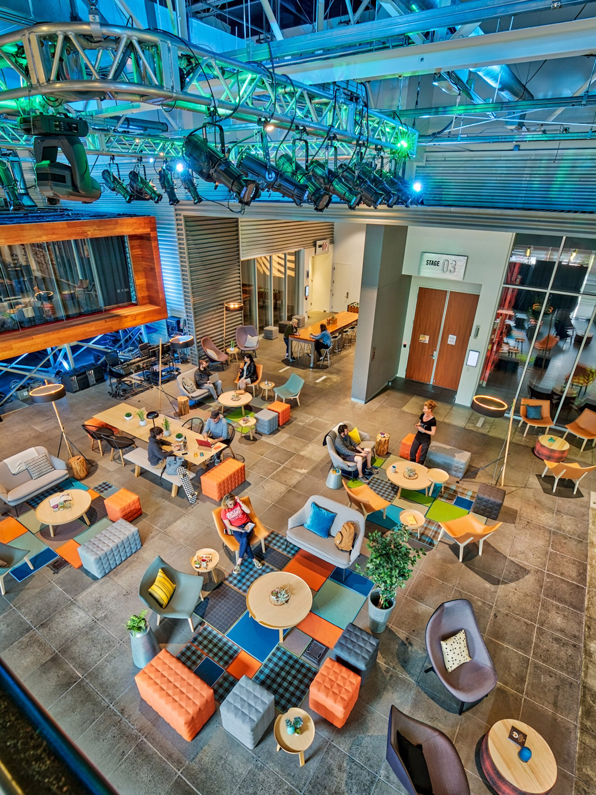 Elevated view of a vibrant, open-plan lounge space featuring modular seating in a mix of citrus and ocean hues. Exposed ceilings with theatrical lighting systems showcase the venue's adaptable nature. Varied table heights and seating styles create a dynamic multi-use area promoting interaction and flexibility.