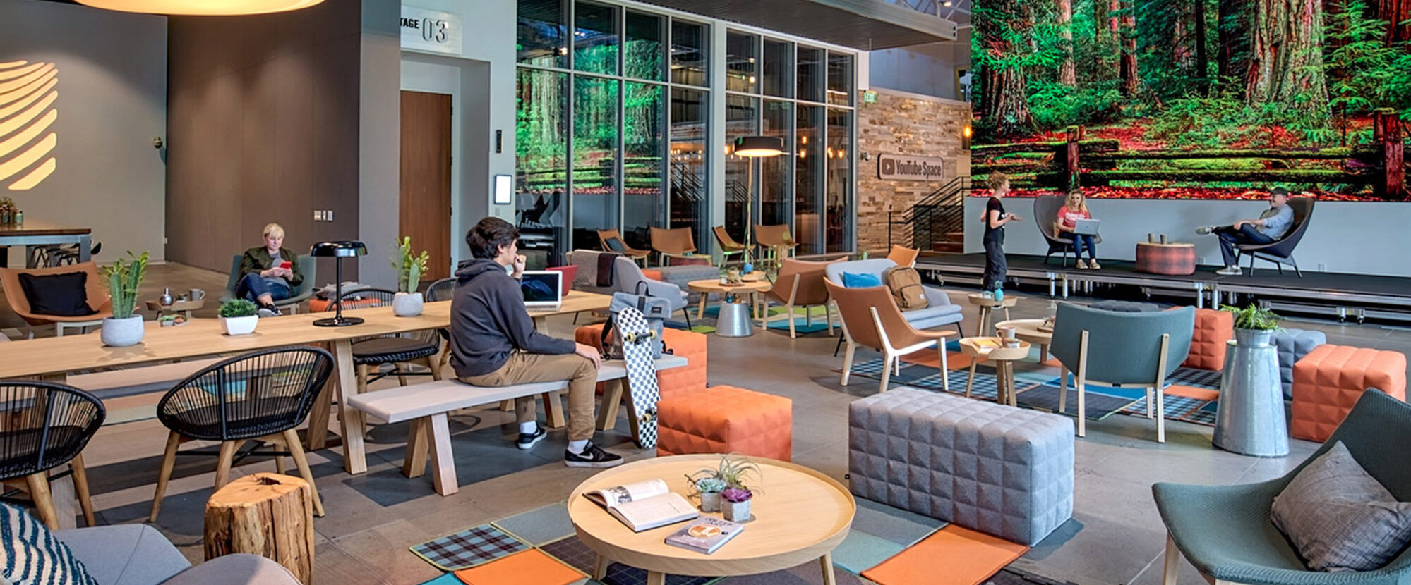 This contemporary workspace balances industrial chic with organic elements, featuring an expansive mural of a forest as a backdrop. Exposed ceiling infrastructure contrasts with warm wooden tables, eclectic seating arrangements, and a mix of geometric and organic patterns. Natural light complements the space's earthy color palette and dynamic layout, promoting a collaborative atmosphere.