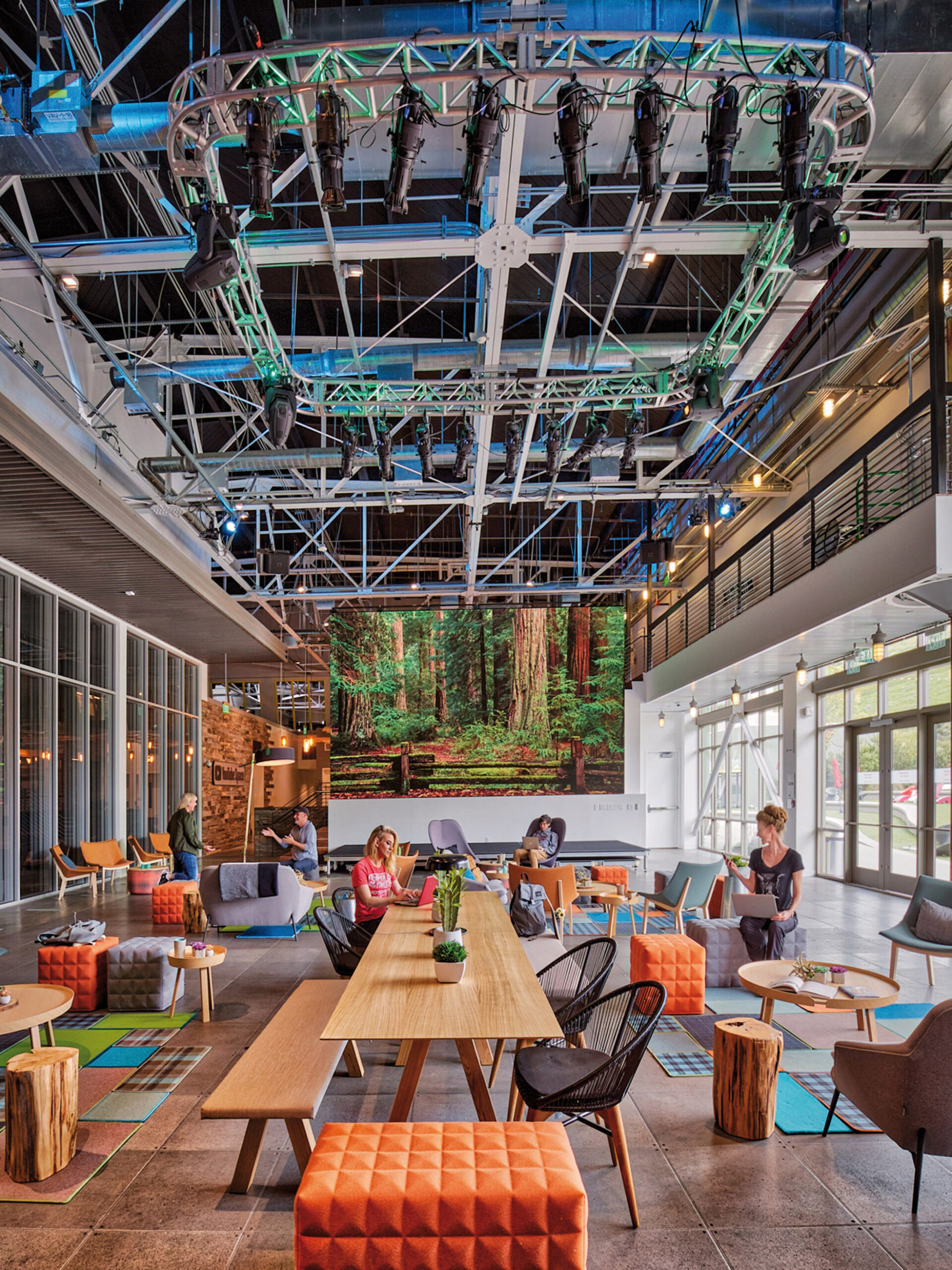 Exposed structural and mechanical elements grace the high ceiling of this vibrant, modern lobby, contributing to its industrial-chic aesthetic. A mix of natural textures and colorful furniture, including wooden tables and eclectic seating options, creates a dynamic, inviting atmosphere. A large digital wall displaying a forest scene adds depth and brings an element of nature indoors, enhancing the biophilic design. Large windows allow for ample natural light