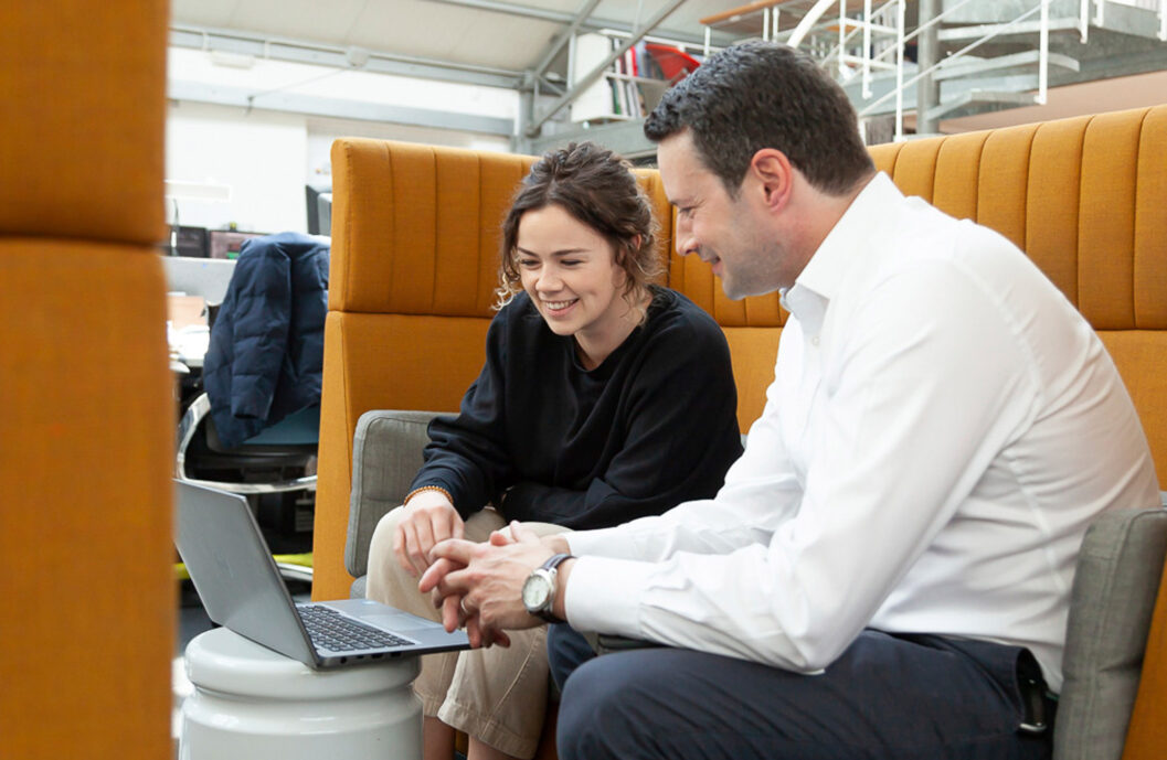 A professional mentor and mentee sitting on an orange couch, engaging in a productive discussion over a laptop. The mentee, a young woman with short hair, smiles as she looks at the screen, while the mentor, a man in a white shirt, points at something of interest on the laptop. The backdrop suggests a bright, open-plan office environment.
