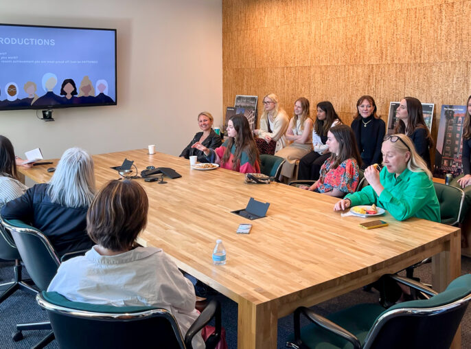 A group of women in a business meeting, engaging in introductions with a slide on the monitor, in a room with a large wooden table and a cork wall, likely celebrating International Women's Day.