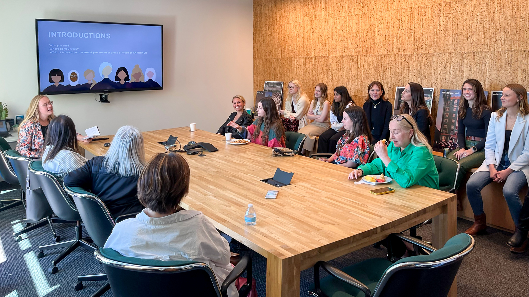 A group of women in a business meeting, engaging in introductions with a slide on the monitor, in a room with a large wooden table and a cork wall, likely celebrating International Women's Day.