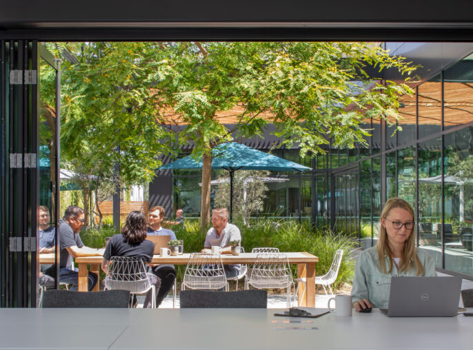 Professionals gather at a modern outdoor meeting space featuring minimalistic furniture and lush greenery, with a transparent glass facade integrating the interior office environment and the vibrant outdoor setting.