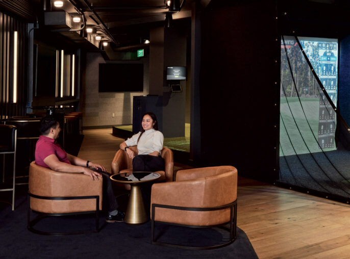 Modern urban-inspired lounge with plush leather seating and industrial-style lighting against a backdrop of dark hues, complemented by a vibrant, full-wall golf simulation screen, offering a dynamic contrast and engaging recreational feature within the space.