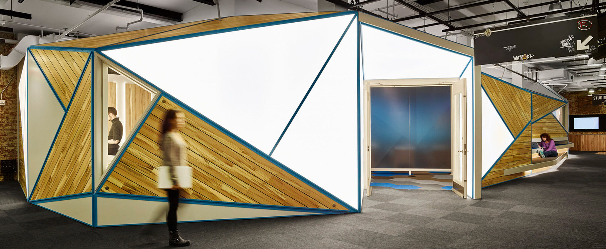 Modern office space featuring angular meeting pods with wooden slats and teal accents. Sleek, exposed ceiling fixtures add an industrial touch above the dynamic workspace layout.
