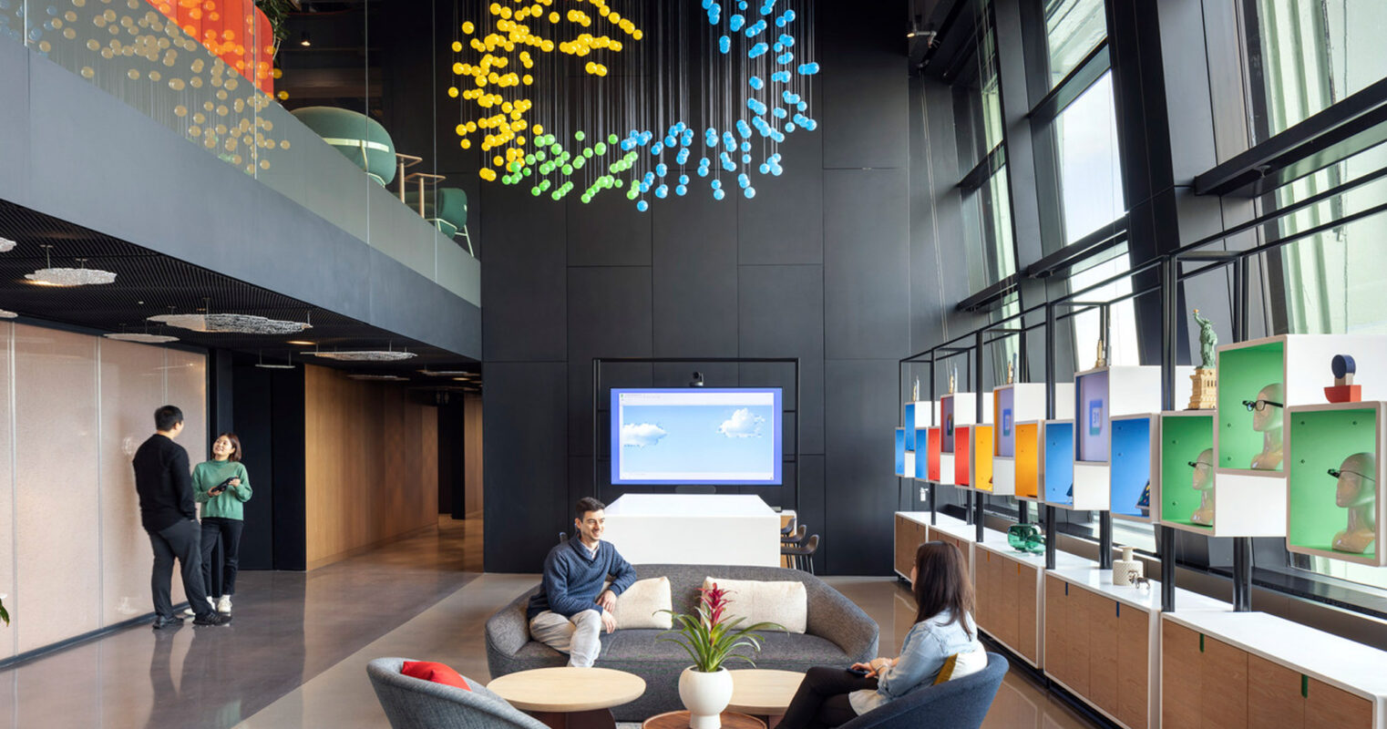 Modern office lobby with eclectic decor, featuring colorful ceiling art installation, mid-century inspired furniture, and glass partition showcasing mounted artwork. An open-concept space fostering collaboration, with individuals engaged in conversation.
