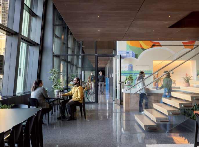 Modern office lobby with polished concrete floors, wood-paneled walls, and full-height windows. Sleek, black communal tables pair with minimalist chairs. A glass-enclosed staircase adds an airy feel, while indoor plants introduce a touch of greenery.