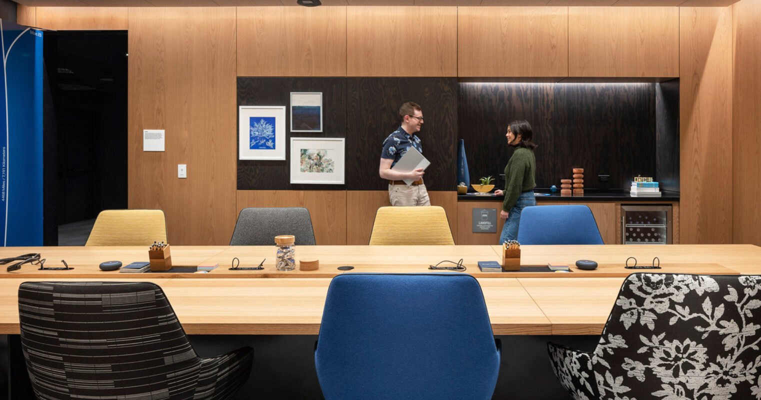 Contemporary office meeting space with warm wooden paneling and integrated shelving. Mustard yellow and royal blue office chairs punctuate the neutral palette. Overhead lighting casts an ambient glow, enhancing the room's inviting atmosphere.