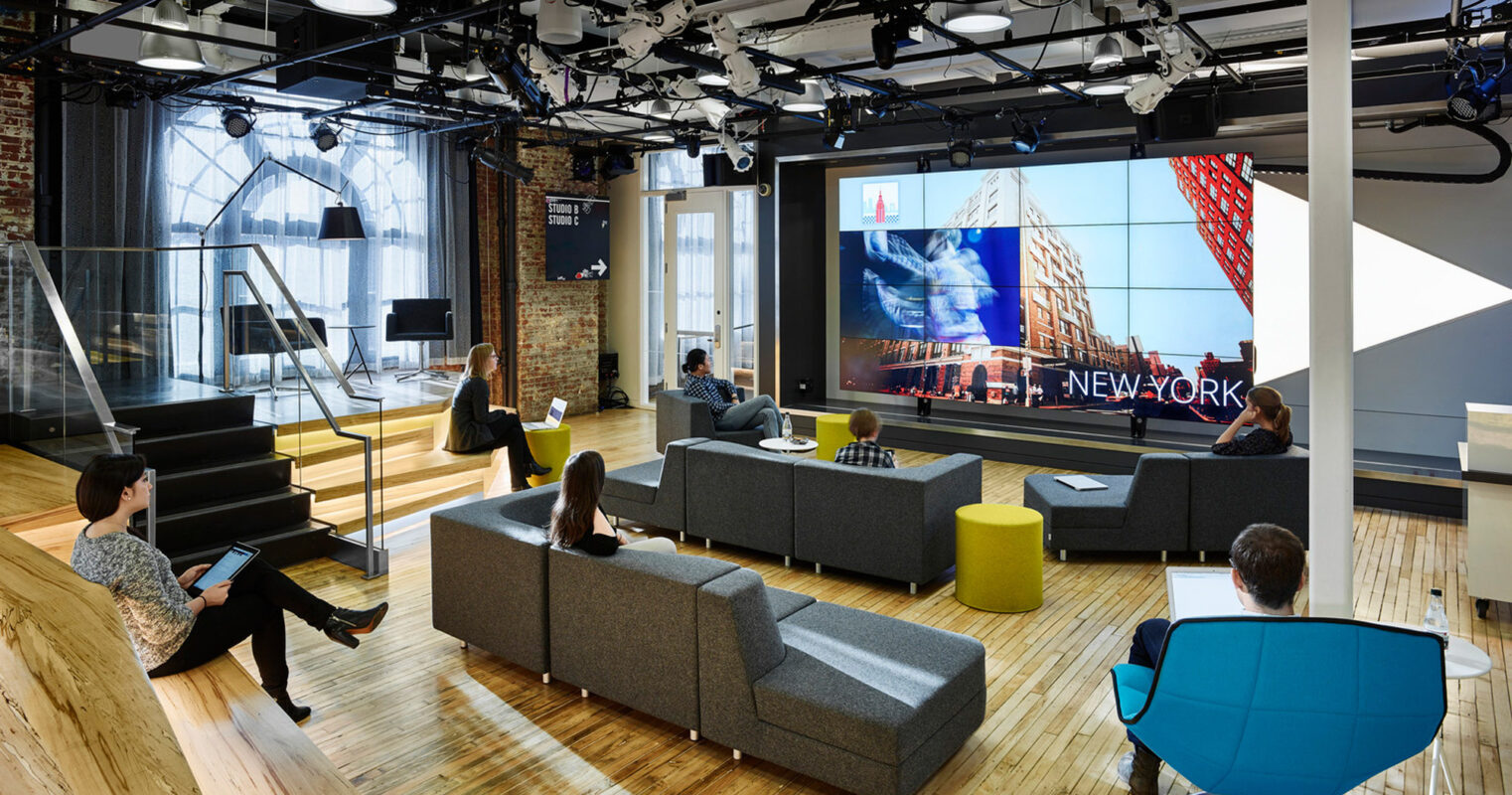 Contemporary office lounge with tiered seating, incorporating a mix of wood floors, exposed brick, industrial ceiling fixtures, and modern furniture, focused around a large video display wall.