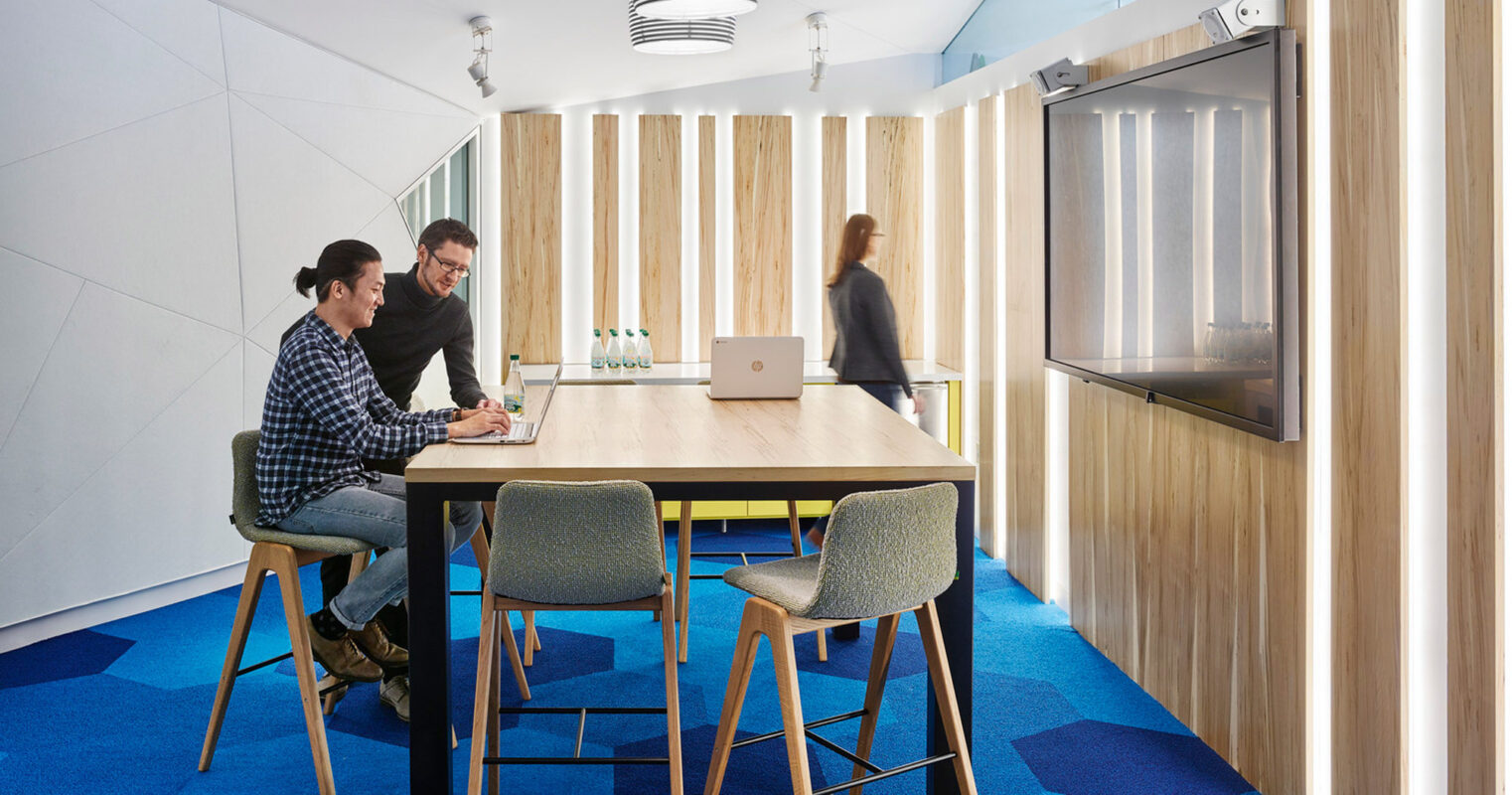 Modern office space with two people collaborating at a table featuring vibrant blue carpet, angular white walls, and a sleek wooden partition. Geometric design elements and a pendant light add to the contemporary aesthetic.
