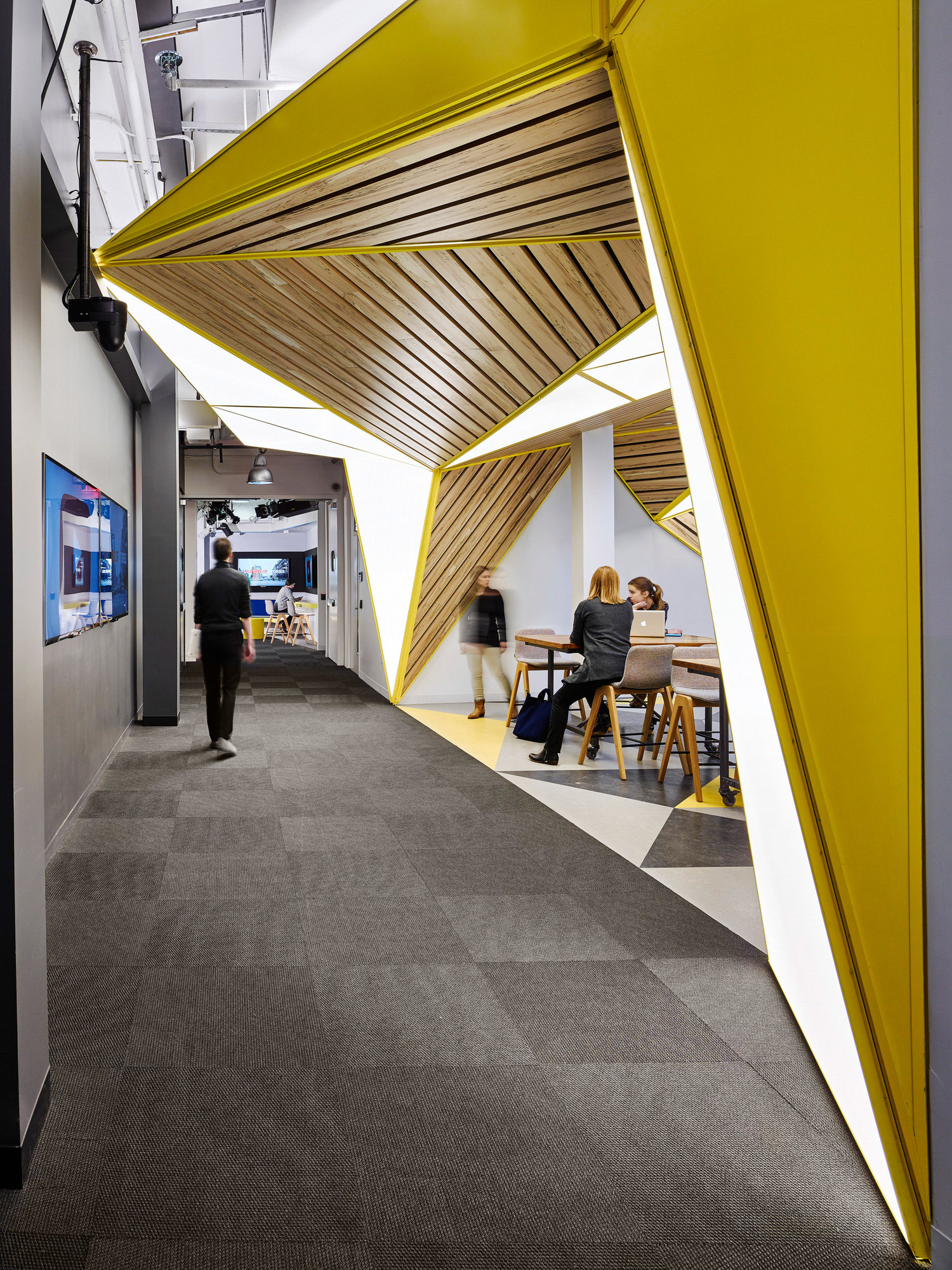 A vibrant modern office space with dynamic geometric forms, featuring a bold yellow triangular entrance that frames the view into a collaborative workspace with wooden elements and informal seating arrangements. The flooring transitions from patterned carpet to sleek wooden finish, enhancing depth and texture.