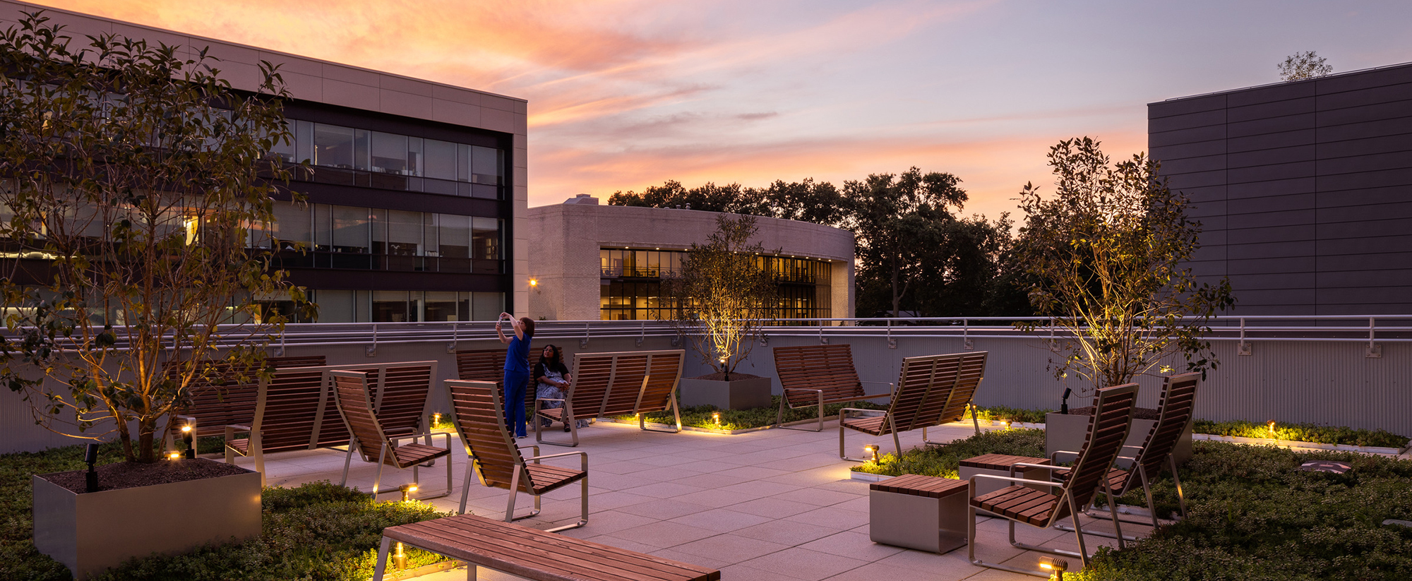 Rooftop terrace at dusk with integrated wooden seating and planters, under-floor lighting, against a backdrop of modern architecture and a vibrant sunset sky.