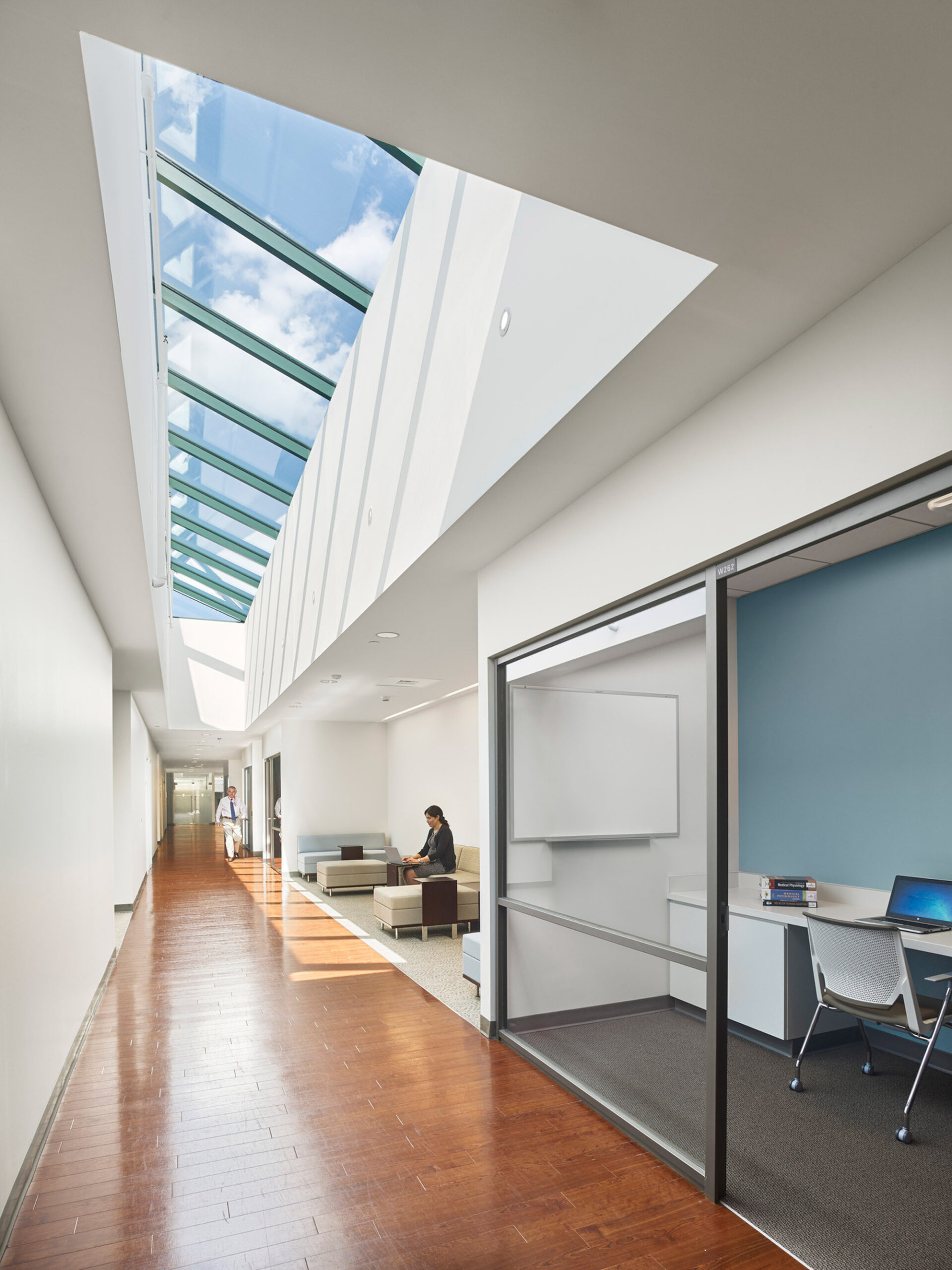 Skylight illuminates a modern hallway with sleek lines and warm wooden floors, juxtaposed against cool-hued walls and minimalist office furniture, accentuating the architectural interplay of light and space.