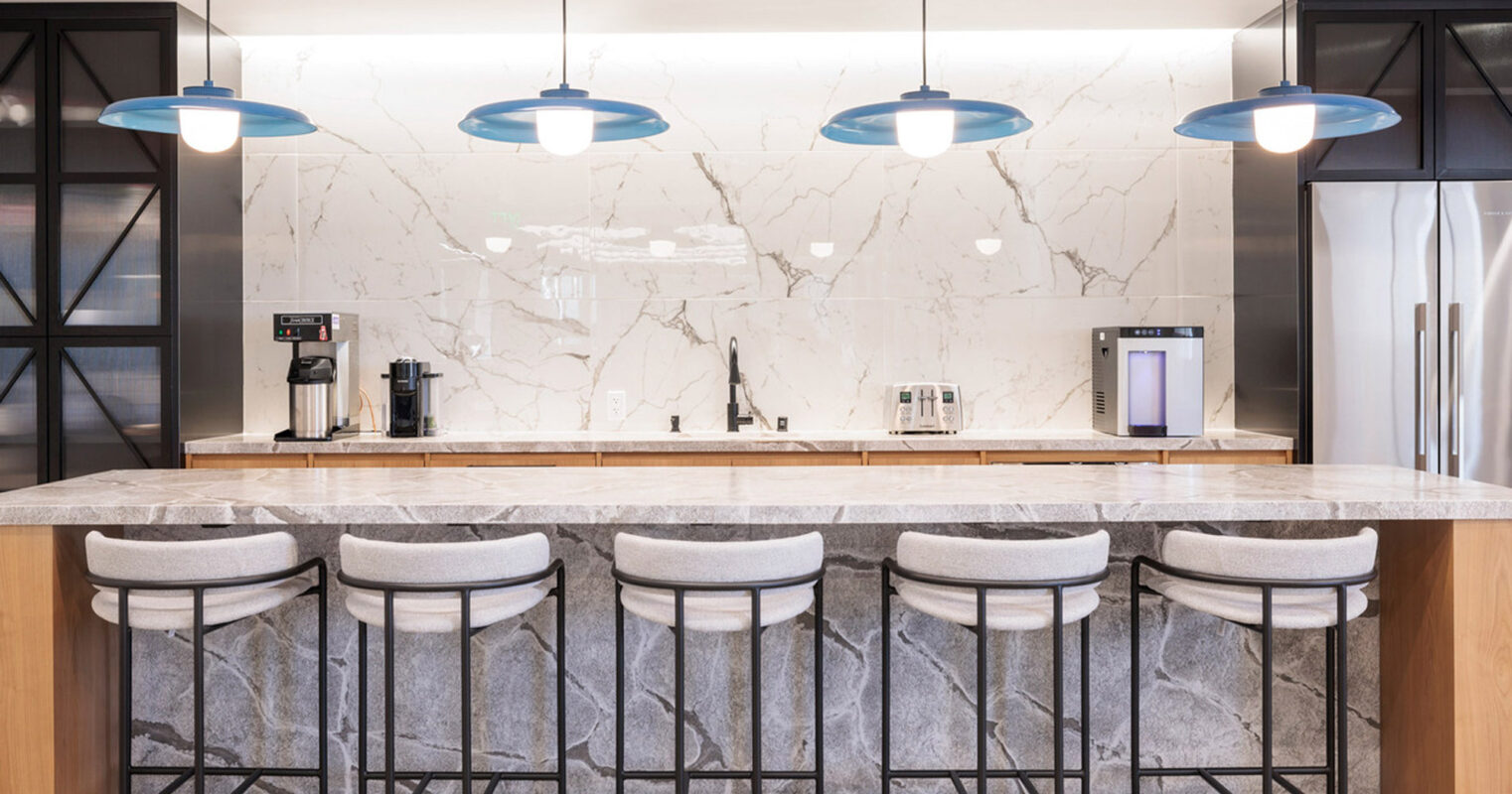 Modern kitchen featuring a white marble backsplash, contrasting with a sleek gray countertop. Overhead, blue pendant lights add a pop of color above the bar-style seating with elegant, gray upholstered stools.