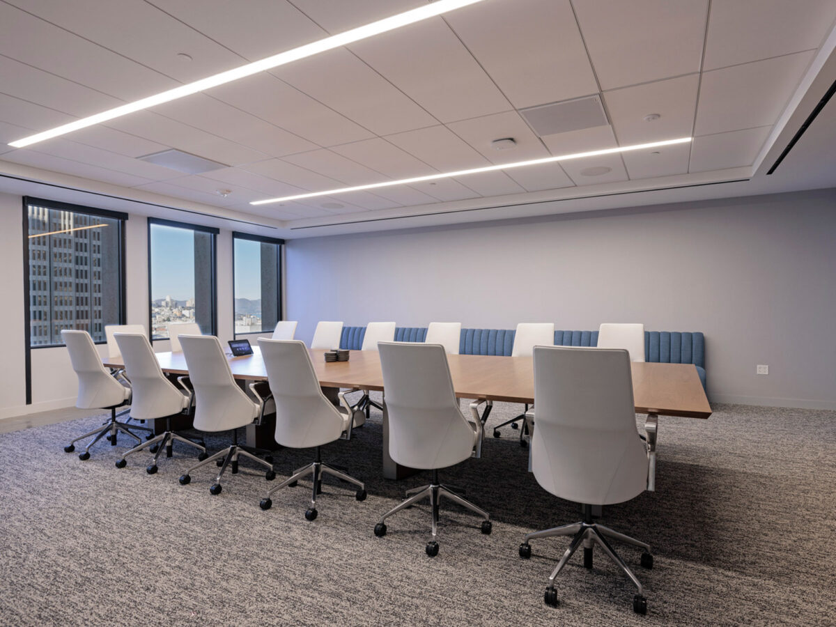 Modern conference room with a sleek, rectangular wooden table and white ergonomic chairs. Recessed LED lighting fixtures provide illumination, complementing the natural light from adjacent windows. Soft gray carpet and a blue upholstered bench along the wall contribute to the room's professional ambiance.