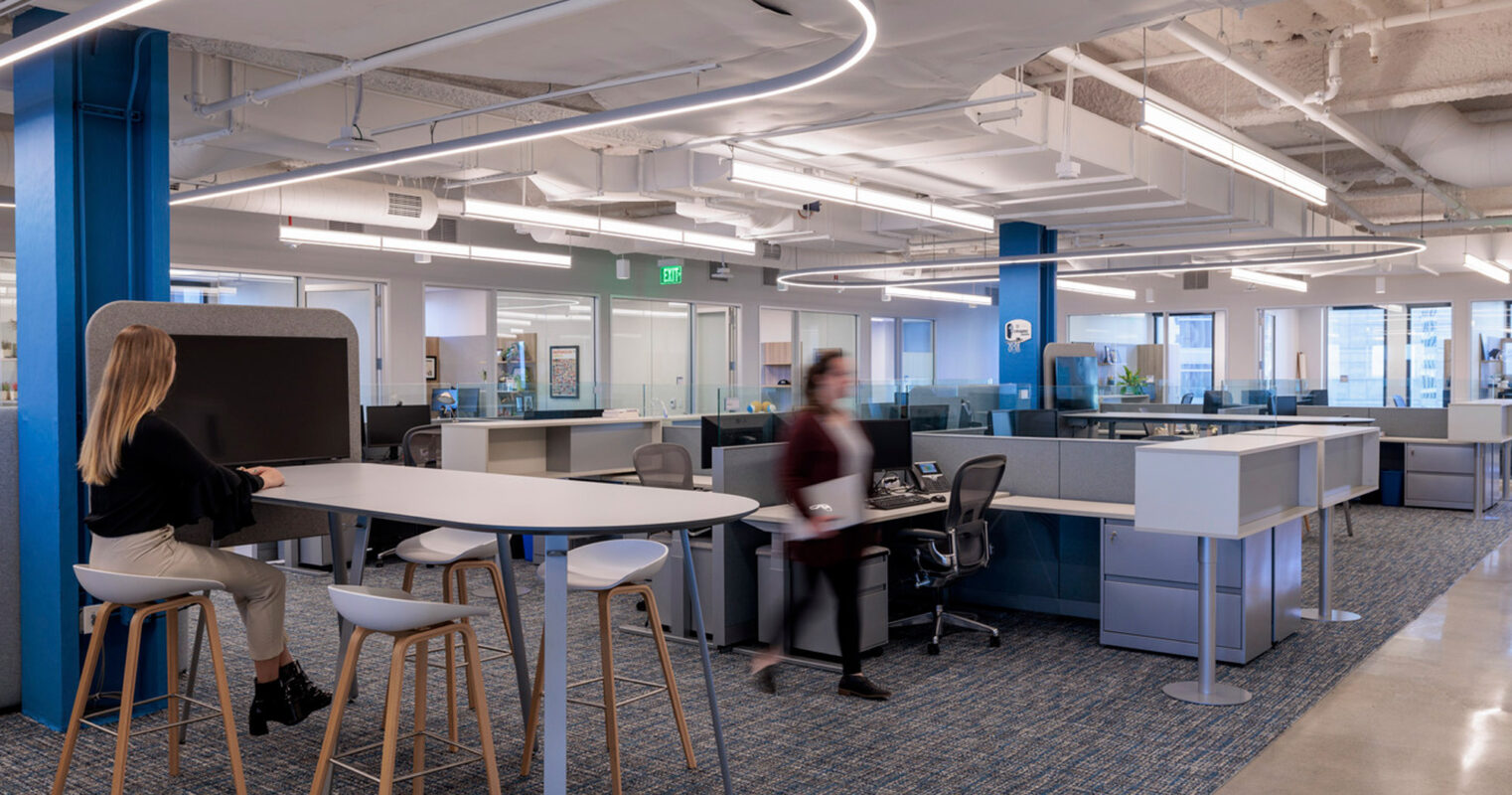 Open-plan office space featuring ergonomic furniture with a combination of standing and traditional desks, accentuated by a dynamic circular overhead lighting structure. The blue structural column adds a pop of color to the soft, neutral palette.