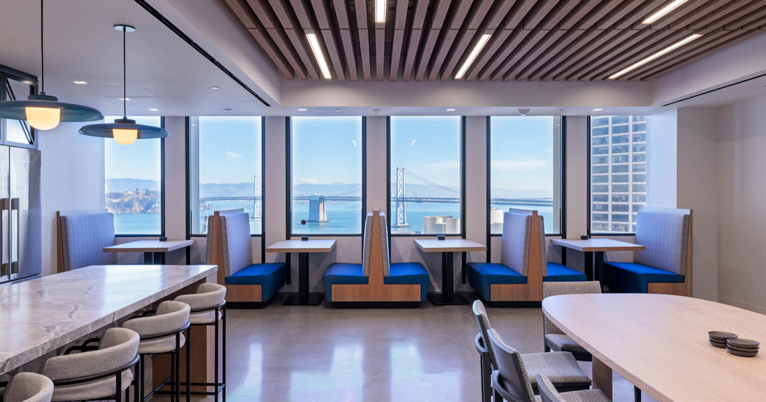 Modern office break room overlooking a bridge through expansive windows, featuring wooden slat ceiling accents, sleek pendant lighting, and two-tone banquette seating. Neutral flooring complements the space's minimalist aesthetic.