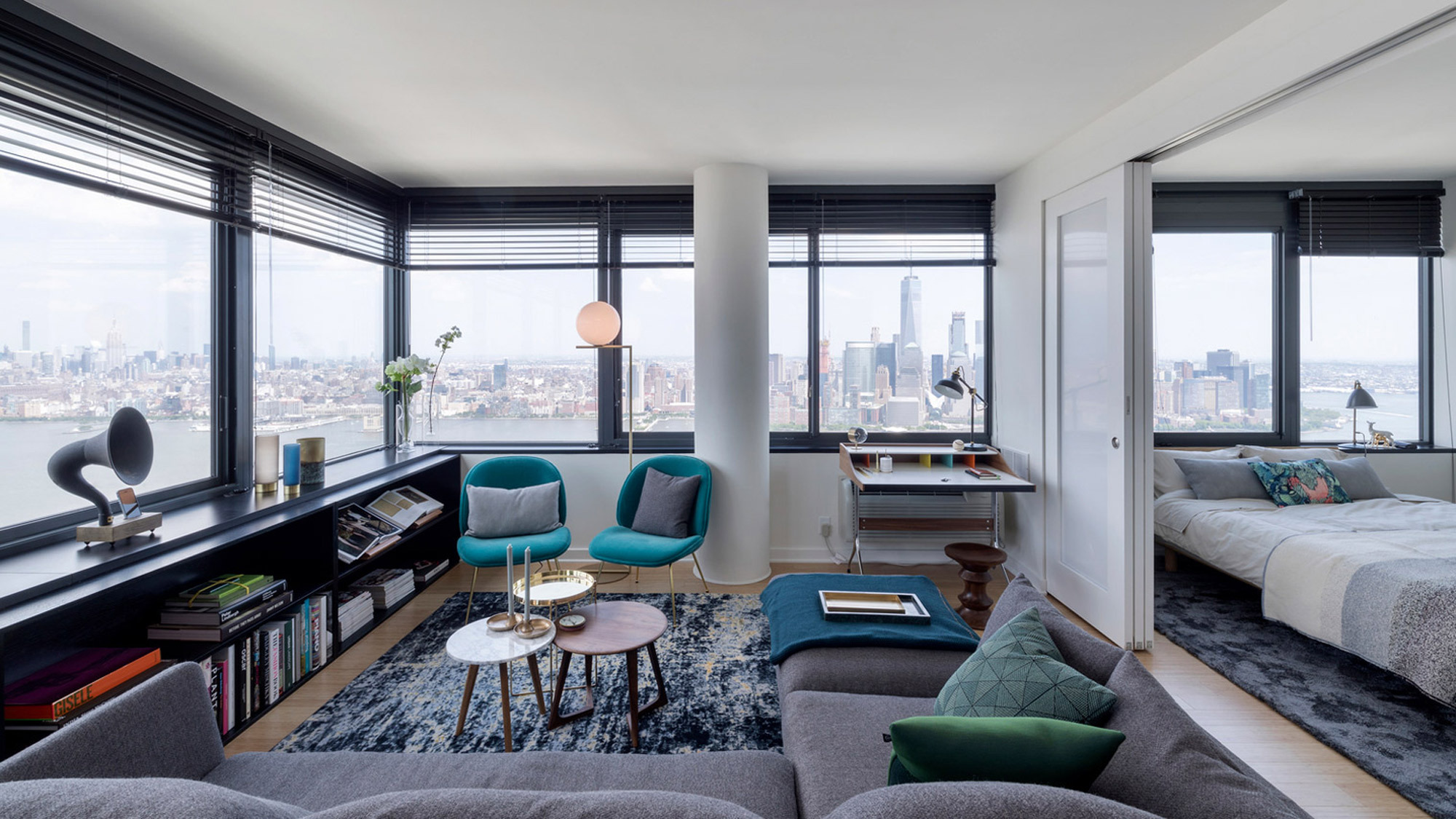 Open-plan living space with floor-to-ceiling windows offering a panoramic city view. Mid-century modern influences seen in teal armchairs and geometric coffee tables. Neutral tones balance the vibrant furniture, and strategic lighting enhances the room's sleek, urban ambiance.