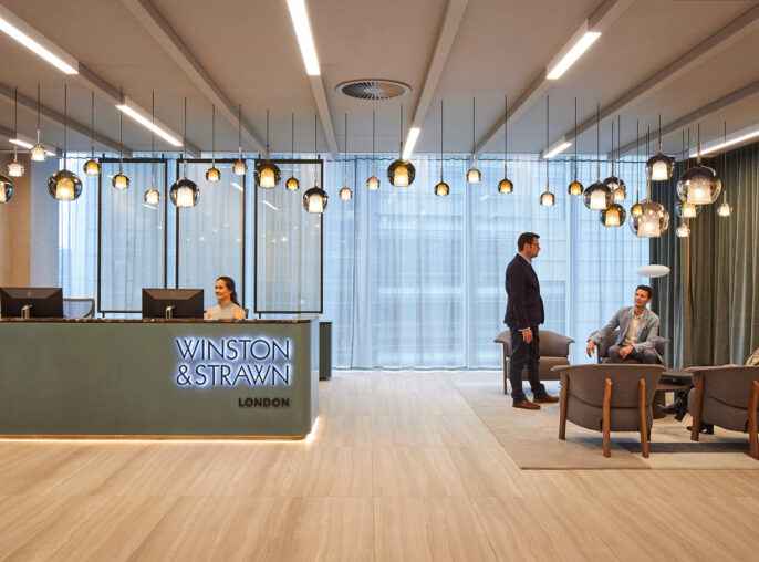 Modern corporate lobby with light wood floors, a sleek reception desk, suspended glass pendant lights, and seated area with neutral-toned furniture. Large windows with sheer curtains provide natural light, complemented by recessed and accent ceiling lighting.