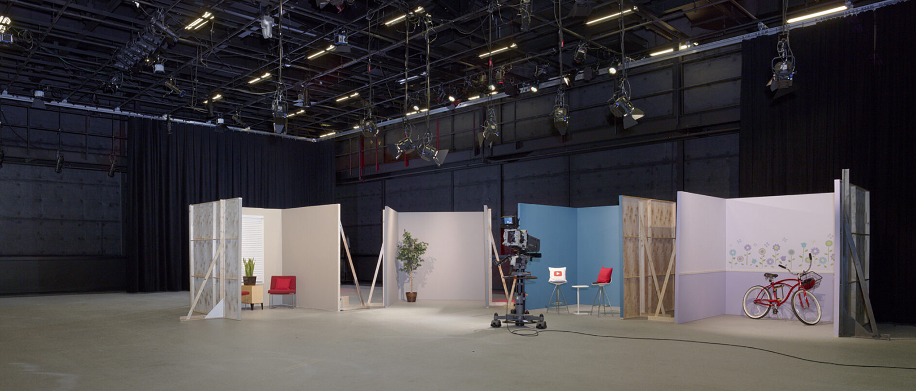 A spacious broadcast studio with high ceilings, equipped with professional lighting rigs and a variety of set pieces including walls, furniture, and props for versatile scene setups.